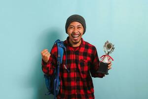 eccitato giovane asiatico uomo alunno indossare un' zaino, Beanie cappello, e rosso plaid flanella camicia, fabbricazione un' sì gesto mentre Tenere un' trofeo e gioia nel risultato, isolato su un' blu sfondo foto