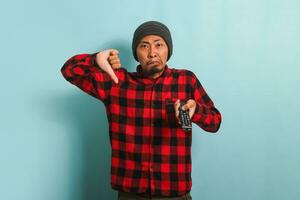 scontento giovane asiatico uomo con un' Beanie cappello e rosso plaid flanella camicia dando un' pollice giù per un' cattivo tv mostrare, isolato su un' blu sfondo foto