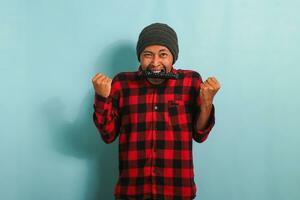 divertente giovane asiatico uomo con un' Beanie cappello e rosso plaid flanella camicia pungente un' tv a distanza, sensazione arrabbiato mentre Guardando un' gli sport gioco su tv, isolato su un' blu sfondo foto