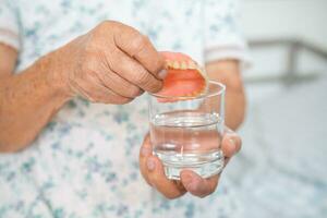 asiatico anziano donna paziente Tenere e lavaggio dentiera nel acqua detersivo bicchiere per bene masticare. foto