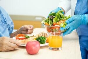 asiatico anziano donna paziente mangiare salmone bistecca prima colazione con verdura salutare cibo mentre seduta e Affamato su letto nel Ospedale. foto