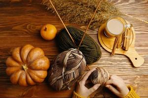 mani in maglione arancione con filato, ferri da maglia, caffè e bastoncini di cannella sul tavolo di legno foto