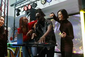 gruppo musicale utilizzando dj stazione e danza su palcoscenico a discoteca raccolta nel discoteca. africano americano frontman giocando whille diverso donne in movimento e coinvolgente folla per festa a vivere concerto foto