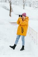 bellissimo ragazza nel un' giallo giacca fotografo prende immagini di neve nel un' inverno parco foto