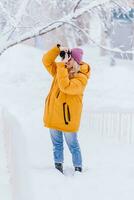 bellissimo ragazza nel un' giallo giacca fotografo prende immagini di neve nel un' inverno parco foto
