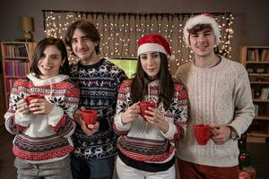 gruppo di quattro giovane amici tostatura con rosso tazze vestito nel rosso cappelli e Natale felpe foto