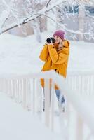 bellissimo ragazza nel un' giallo giacca fotografo prende immagini di neve nel un' inverno parco foto