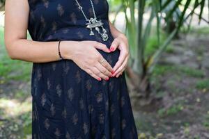 un' incinta indiano signora pose per all'aperto gravidanza sparare e mani su gonfiarsi, indiano incinta donna mette sua mano su sua stomaco con un' maternità vestito a società parco, incinta al di fuori maternità sparare foto