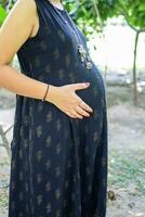 un' incinta indiano signora pose per all'aperto gravidanza sparare e mani su gonfiarsi, indiano incinta donna mette sua mano su sua stomaco con un' maternità vestito a società parco, incinta al di fuori maternità sparare foto