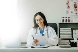 fiducioso giovane asiatico femmina medico nel bianca medico uniforme sedersi a scrivania Lavorando su computer. sorridente uso il computer portatile Scrivi nel medico rivista foto