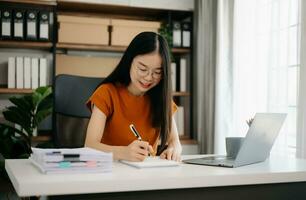 femmina studenti Nota a partire dal il libri a il asiatico ragazza biblioteca seduta a il tavolo utilizzando il computer portatile computer e tavoletta per ricerca un in linea informazioni. foto