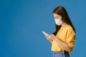 la giovane ragazza asiatica indossa la maschera facciale utilizzando il telefono cellulare su sfondo blu. foto