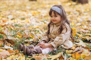 bambina in un cappotto beige seduto tra le foglie nel parco autunnale. foto