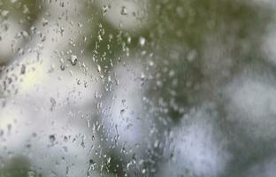 un' foto di pioggia gocce su il finestra bicchiere con un' sfocato Visualizza di il fioritura verde alberi. astratto Immagine mostrando nuvoloso e piovoso tempo metereologico condizioni