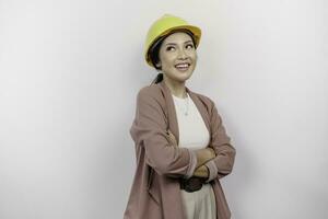 un' fiducioso sorridente asiatico donna dipendente indossare sicurezza casco in piedi con braccia piegato e guardare a il telecamera isolato al di sopra di bianca sfondo foto