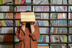 ritratto di asiatico hijab donna copertura sua testa con un' mucchio di libri nel davanti di biblioteca scaffale. musulmano ragazza lettura un' libro. concetto di alfabetizzazione e conoscenza foto