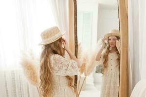 un' bellissimo adolescenziale ragazza con lungo capelli le misure un' cannuccia cappello nel davanti di un' specchio. autoammirazione di un' bionda. foto