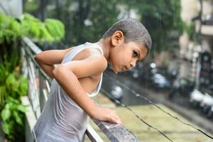 poco ragazzo giocando nel estate pioggia nel Casa balcone, indiano inteligente ragazzo giocando con pioggia gocce durante monsone piovoso stagione, ragazzo giocando nel pioggia foto