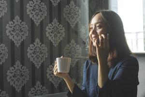 donna asiatica sorridente rilassata che beve caffè mentre chiama per lavoro