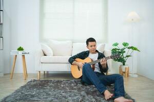 asiatico uomo giocando chitarra seduta su divano divano o pavimento nel il vivente camera a casa. asiatico uomo scrittura canzone mentre giocando chitarra a casa. comporre canzone musica concetto. foto