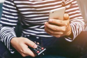 il giovane che utilizza il telecomando della televisione e lo smartphone mobile è seduto su un divano. acceso o spento tv. foto