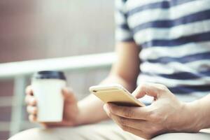 giovane maschio utilizzando su cellula Telefono In piedi nel Guardando Messaggio su mobile Telefono durante rompere mano Tenere carta tazza di prendere lontano potabile caffè caldo su il mattina nel pubblico parco. foto