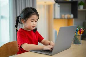 asiatico bambino ragazza indossare un' rosso maglietta uso il computer portatile e studia in linea su legna tavolo scrivania nel vivente camera a casa. formazione scolastica apprendimento in linea a partire dal casa concetto. foto