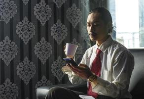 uomo asiatico rilassato che beve caffè mentre usa un telefono cellulare per lavoro