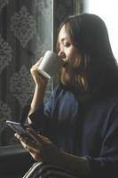 donna asiatica rilassata che beve caffè mentre usa un telefono cellulare per lavoro
