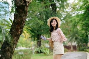 ritratto di asiatico giovane donna viaggiatore con tessitura cappello e cestino contento Sorridi su verde pubblico parco natura sfondo. viaggio viaggio stile di vita, mondo viaggio esploratore o Asia estate turismo concetto. foto