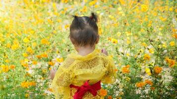 poco carino ragazza indossare giallo balinese vestito giocando nel giallo e bianca fiore giardino foto