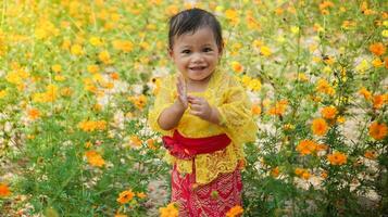 poco carino ragazza indossare giallo balinese vestito giocando nel giallo e bianca fiore giardino foto