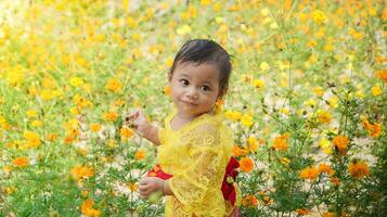 poco carino ragazza indossare giallo balinese vestito giocando nel giallo e bianca fiore giardino foto