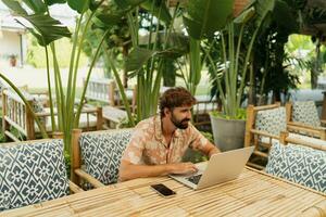 bello uomo con barba utilizzando giro superiore e mobile Telefono , seduta nel all'aperto bar con tropicale interno. foto