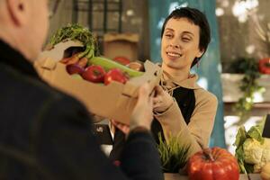 giovane piacevole donna dando scatola pieno di fresco biologico frutta e verdure per cliente mentre Lavorando a Locale agricoltori mercato. femmina contadino vendita salutare localmente cresciuto azienda agricola cibo foto