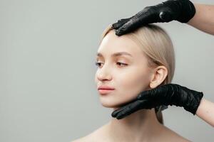 il mani di un' cosmetologo chirurgo nel nero guanti toccare un' donna di viso isolato su un' leggero sfondo, un' bellissimo ragazza con pulito pelle senza trucco controlli il pelle di il viso a un' cosmetologo foto