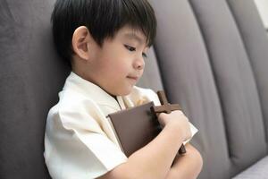 poco asiatico ragazzo prega Tenere un' attraversare e un' religioso prenotare, cristiano concetto foto