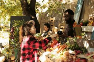 africano americano tipo venditore in piedi dietro a stalla vendita fresco biologico di stagione verdure per femmina cliente a Locale agricoltori mercato. giovane donna acquisto naturale homegrown produrre foto