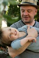 padre detiene contento poco figlio nel il suo braccia nel primavera giardino. padre giorno, famiglia affetto concetto. foto