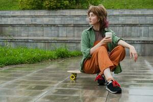 gen z pattinatore ragazza seduta su skateboard con caffè carta contenitore nel sua mano. foto