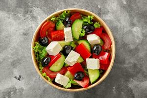 horiatiki insalata con tritato la verdura, lattuga, feta formaggio e nero olive nel carta ciotola su grigio pietra superficie foto