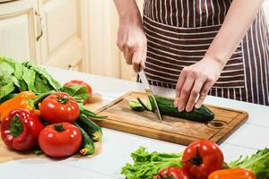 giovane donna cucinando nel il cucina a casa. un' donna tagli un' cetriolo e verdure con un' coltello. foto