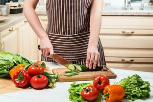 giovane donna cucinando nel il cucina a casa. un' donna tagli un' cetriolo e verdure con un' coltello. foto