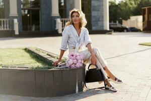 elegante bellissimo allegro biondo donna nel elegante casuale attrezzatura , Tenere rosa boquet con pioni fiori. in posa su il strada. foto