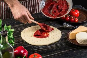 un' maschio mano diffusione pomodoro puré su un' Pizza base con cucchiaio su un vecchio di legno sfondo foto