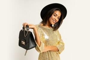 studio foto di bellissimo nero donna nel d'oro festa vestito posa su bianca sfondo. indossare elegante cappello e orecchini. Tenere pelle Borsa.