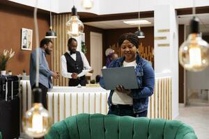 contento del Millennio africano americano donna in piedi nel Hotel atrio guardare a il computer portatile schermo, fabbricazione video chiamata mentre in viaggio solo. femmina libero professionista godendo a distanza lavoro, Lavorando remoto su vacanza foto