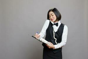 sorridente asiatico cameriera segreteria telefono fisso Telefono chiamata e scrittura su clipboard. ristorante amichevole donna dipendente nel uniforme parlando con cliente su telefono e gestione ordine foto