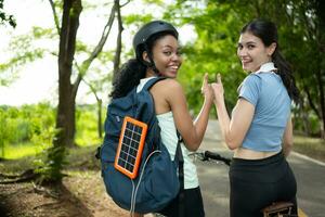 tutti e due di giovane donne nel abbigliamento sportivo equitazione un' bicicletta nel il parco, trasportare un' zaino con un' solare pannello per caricare smartphone. foto