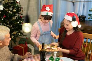 contento famiglia festeggiare Natale insieme a casa. allegro genitori e bambini nel Santa cappello dando cestino con biscotto per loro bambini. foto